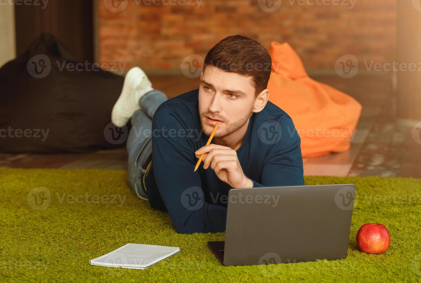 Thoughtful Guy Using Laptop Thinking Creating Startup Lying On Floor In Modern Coworking Workspace. Creative Job And Freelance Lifestyle photo