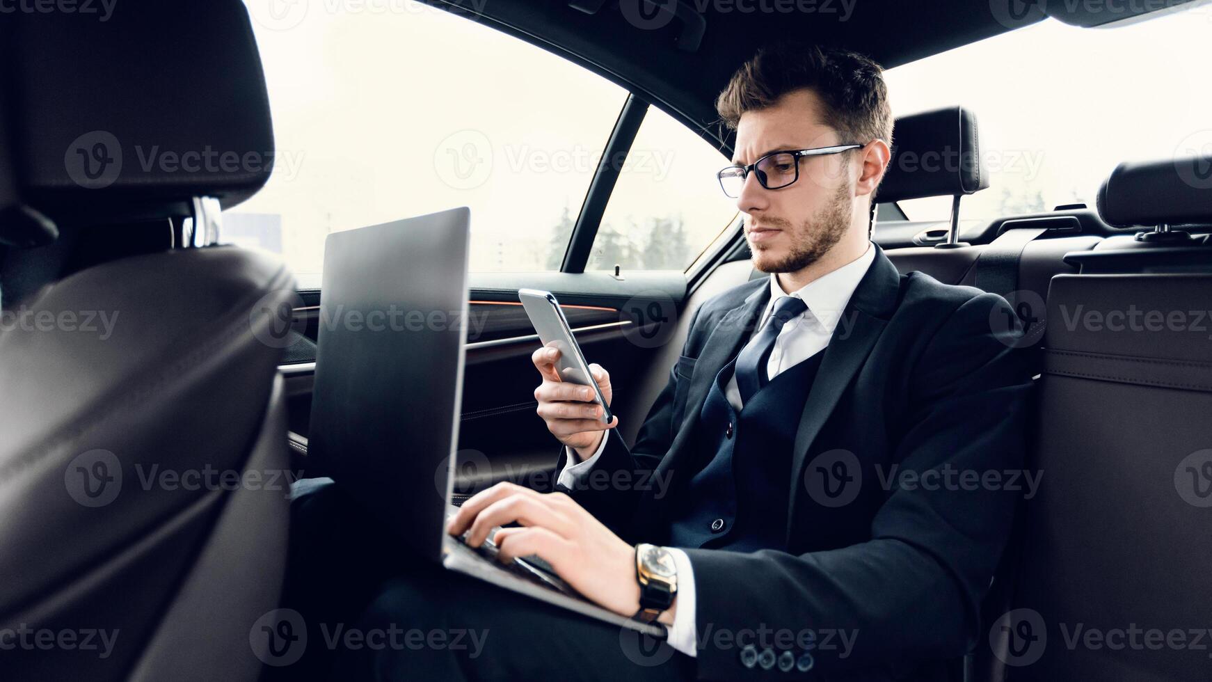 Multitasking. Bearded businessman wearing suit and glasses sitting in back seat of car, using smart phone and laptop photo