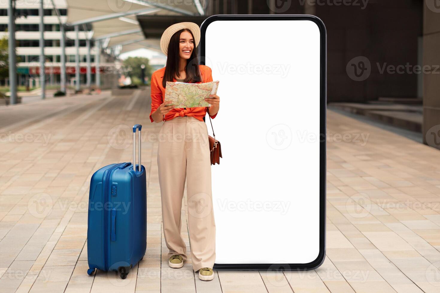 A traveler with a bright outfit holds a map while standing next to a large blank screen, ready to explore the city. A blue suitcase rests beside her in a lively urban area. photo