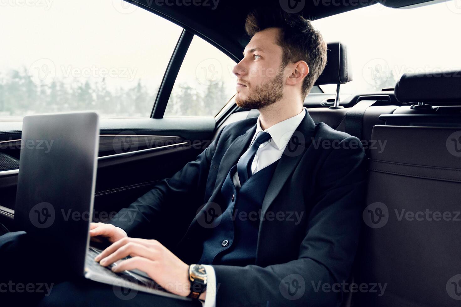 Always In Touch. Handsome businessman working on laptop, sitting in backseat of luxury car, looking out of window photo
