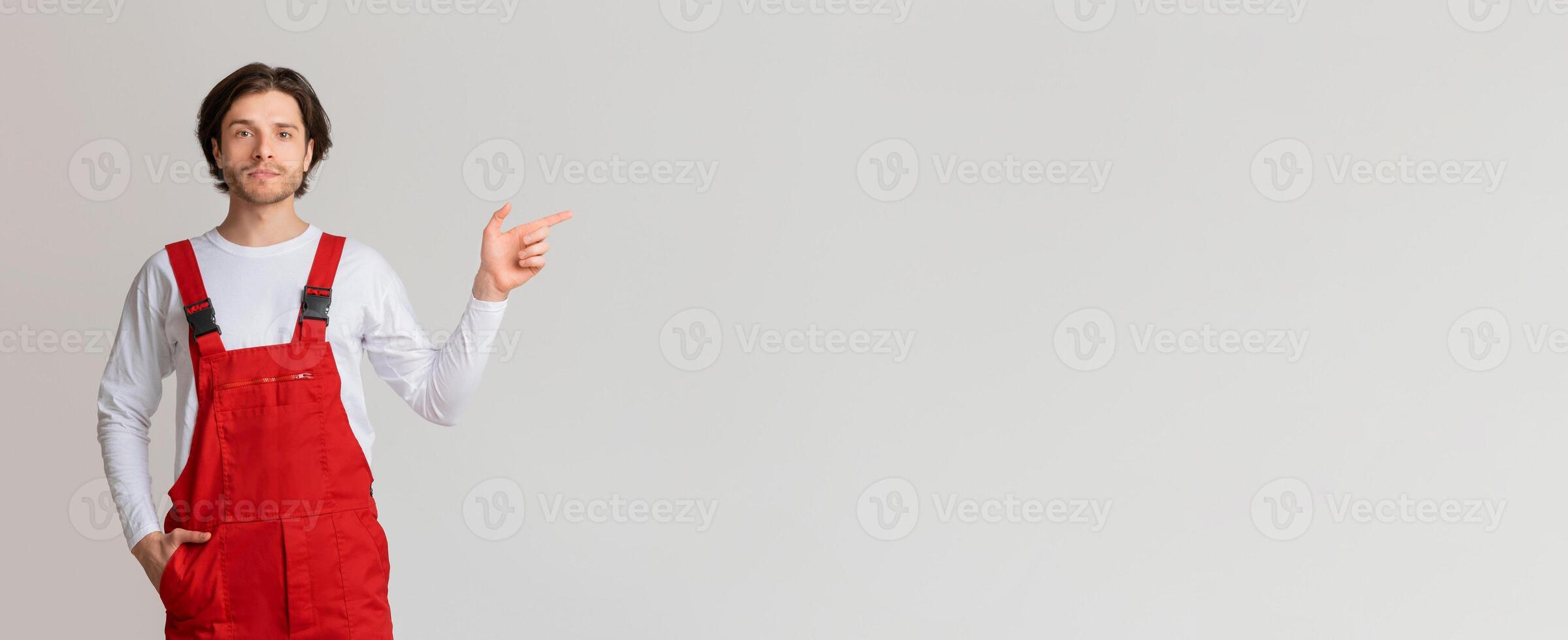 Young confident workman in uniform pointing aside at copy space on light background, panorama photo