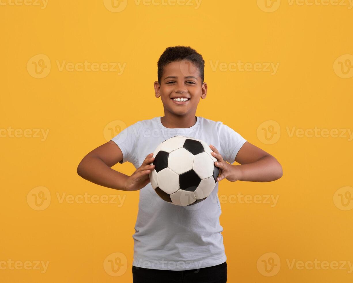 Cheerful black school boy holding soccer ball and smiling at camera, playing football, yellow studio background, copy space. Sport, leisure, entertainment, hobby for kids concept photo