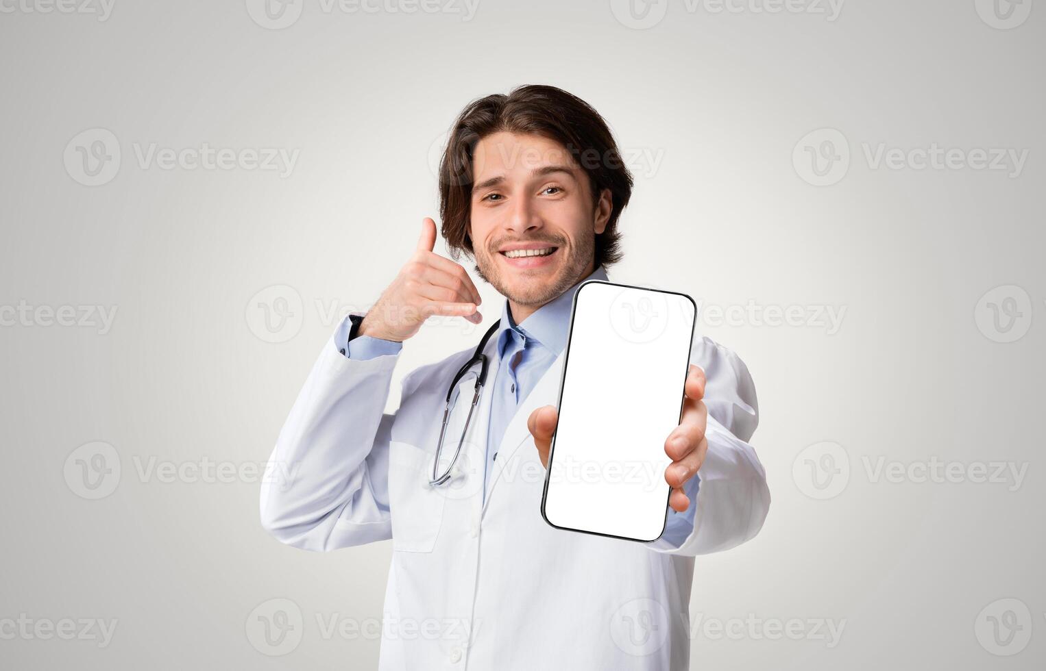 Positive medical specialist pointing at camera and showing call gesture over white studio background, empty space photo