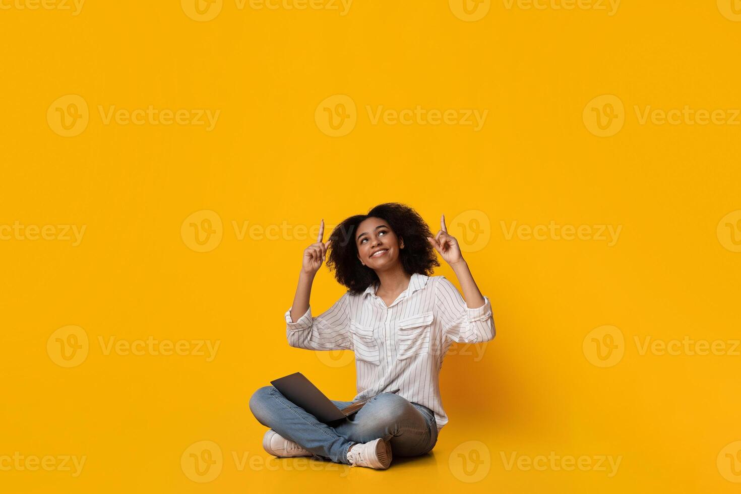 Great Offer. Excited black woman with laptop on lap sitting on floor and pointing up at copy space above her head over yellow studio background photo