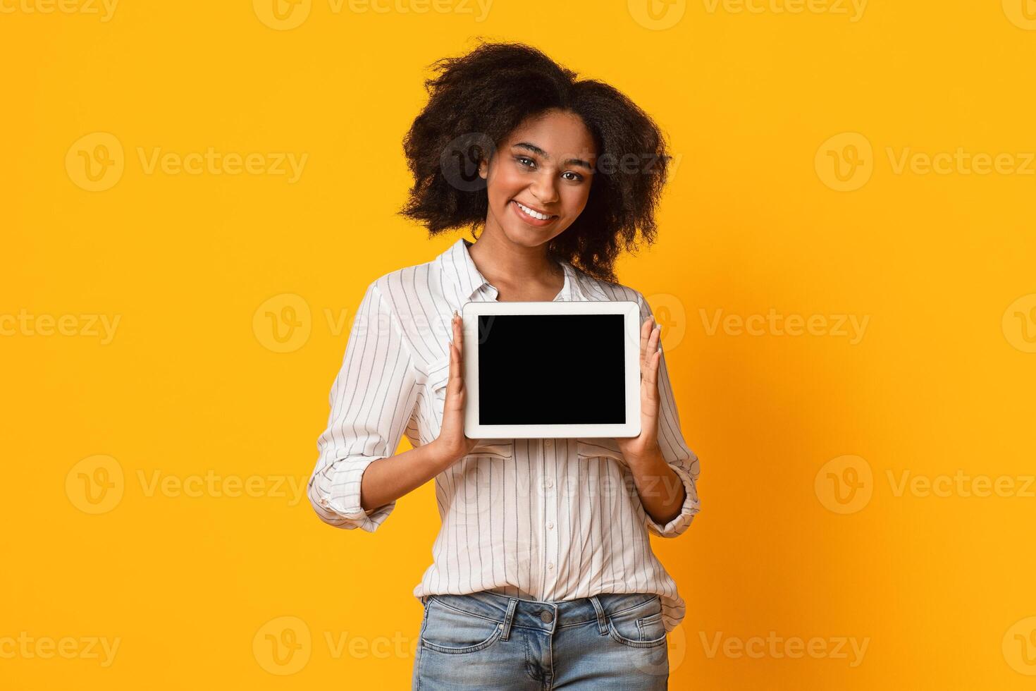 Cheerful Black Girl Showing Digital Tablet With Black Blank Screen, Holding Device In Her Hands And Sniling At Camera, Yellow Background, Mockup photo