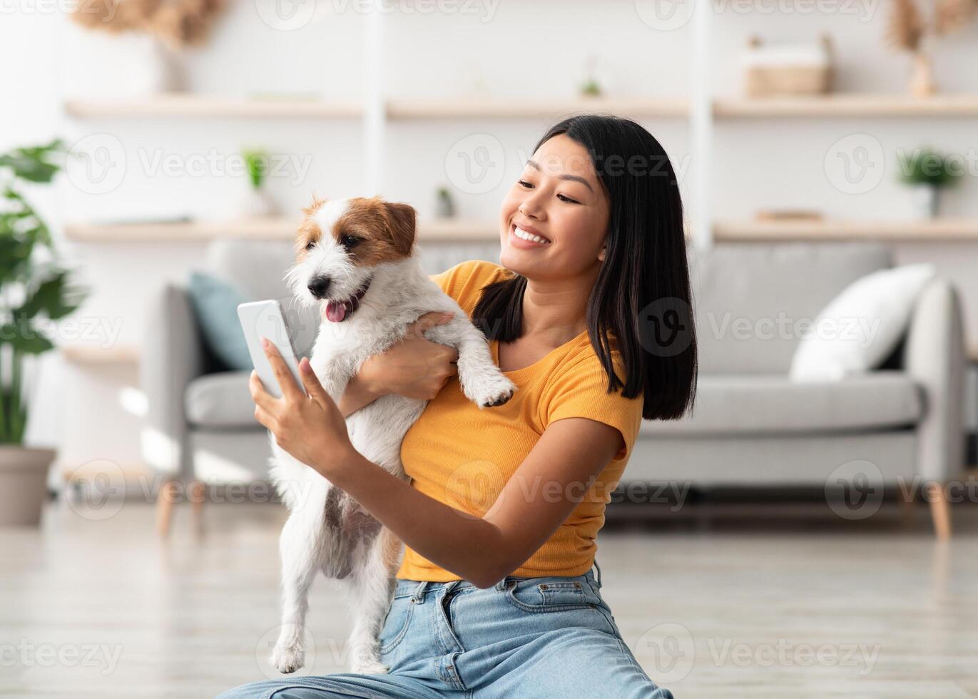 Happy young asian woman in casual sitting on floor in living room, taking selfie with her playful cute dog jack russel terrier puppy while playing together at home, using mobile phone, copy space photo