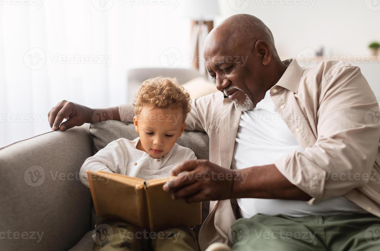 African American Grandpa And Grandson Reading A Book Spending Time Together Sitting On Couch At Home On Weekend. Senior Grandfather Teaching Grandchild To Read Indoors. Knowledge, Family Leisure photo