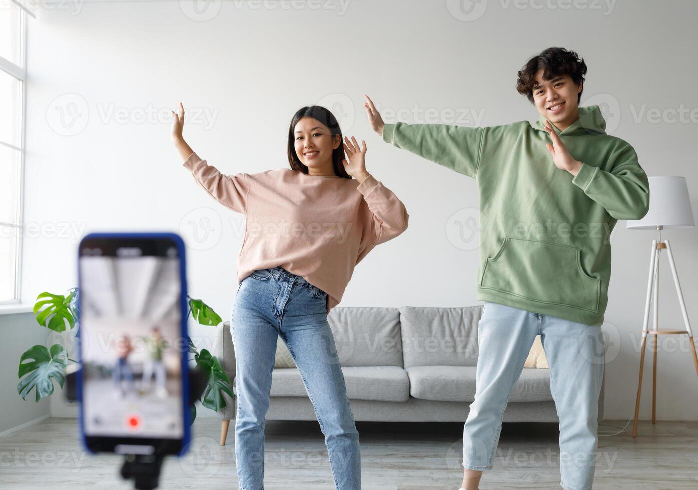 Young Asian couple recording content, dancing on smartphone camera at home. Positive millennial boyfriend and girlfriend filming for social media, using mobile device. Vlogging concept photo
