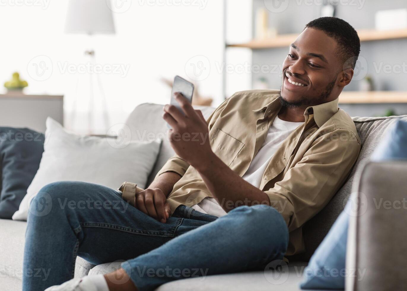 Side View Of African American Guy Using Smartphone Browsing Internet Sitting On Couch At Home. Black Man Texting On Cellphone Or Using New Application On Mobile Phone Indoors photo