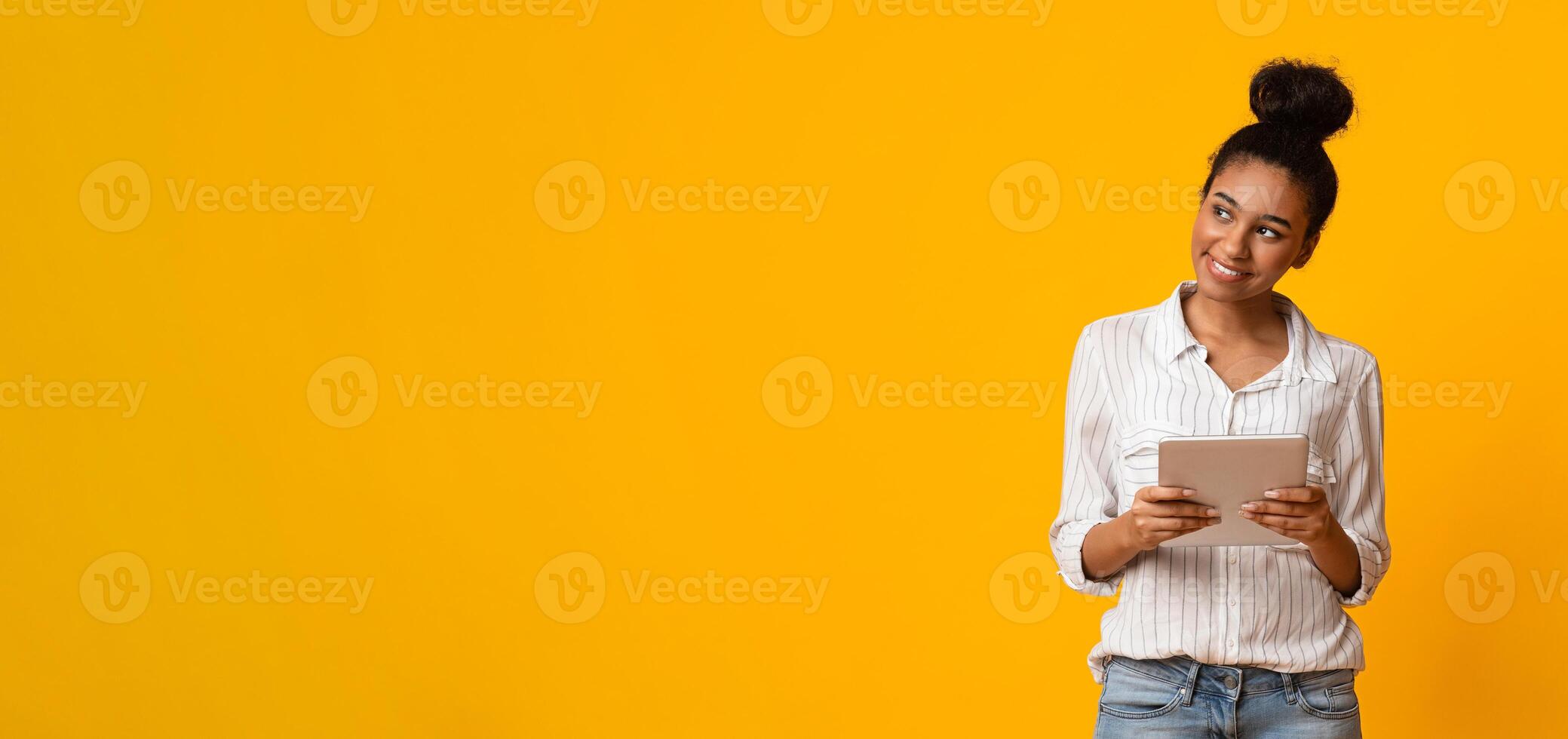 Interested black woman with digital tablet in hands looking aside at copy space on yellow background, panorama photo