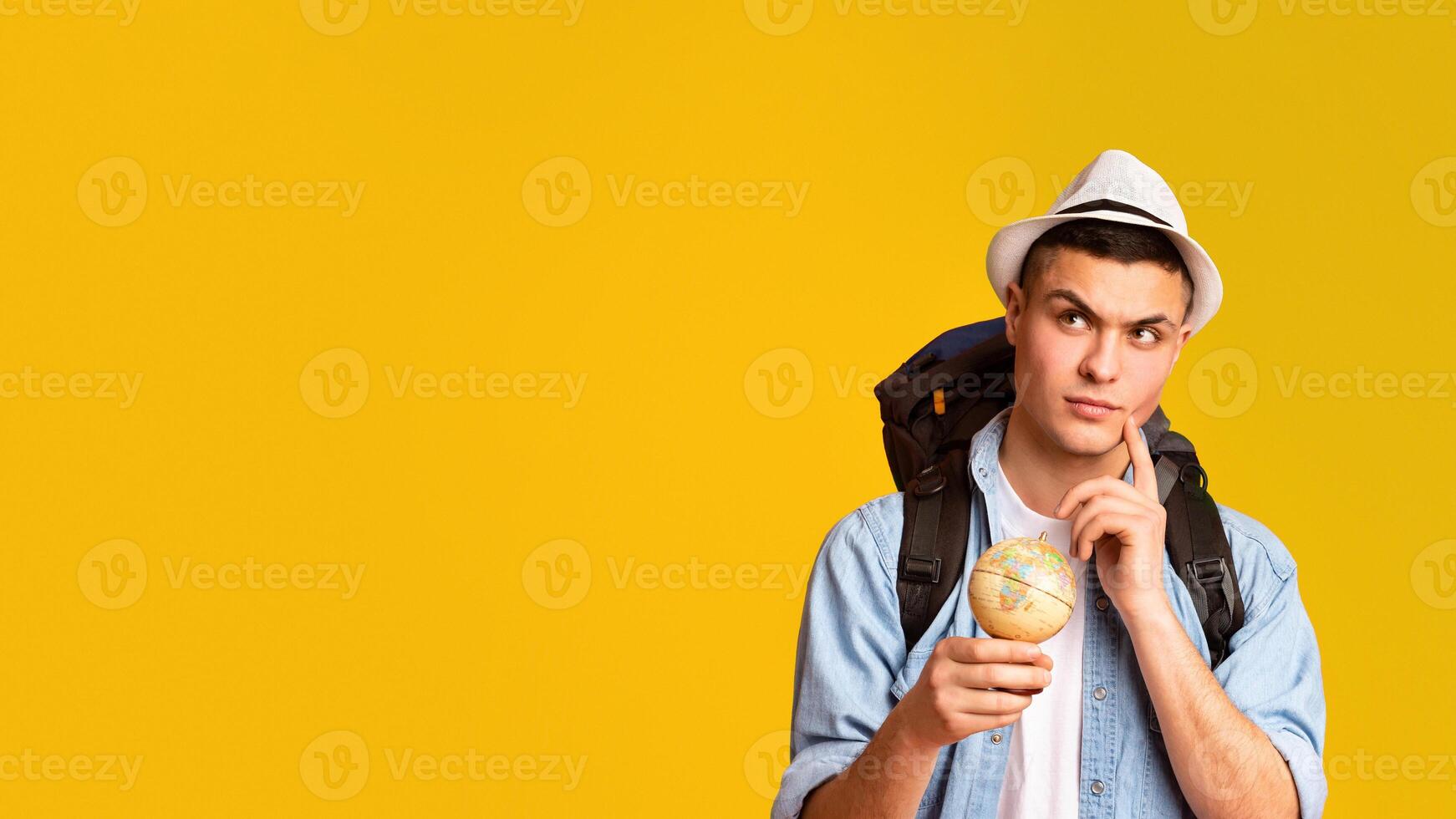 Dont't know where to go. Thoughtful traveler with big backpack and globe over yellow studio background, panorama with free space photo