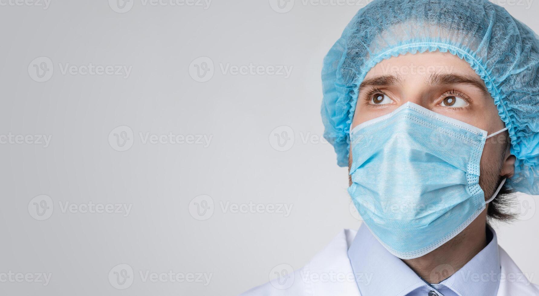 Coronavirus Fight. Confident male doctor in medical mask and hair net looking at copy space over light background, panorama photo