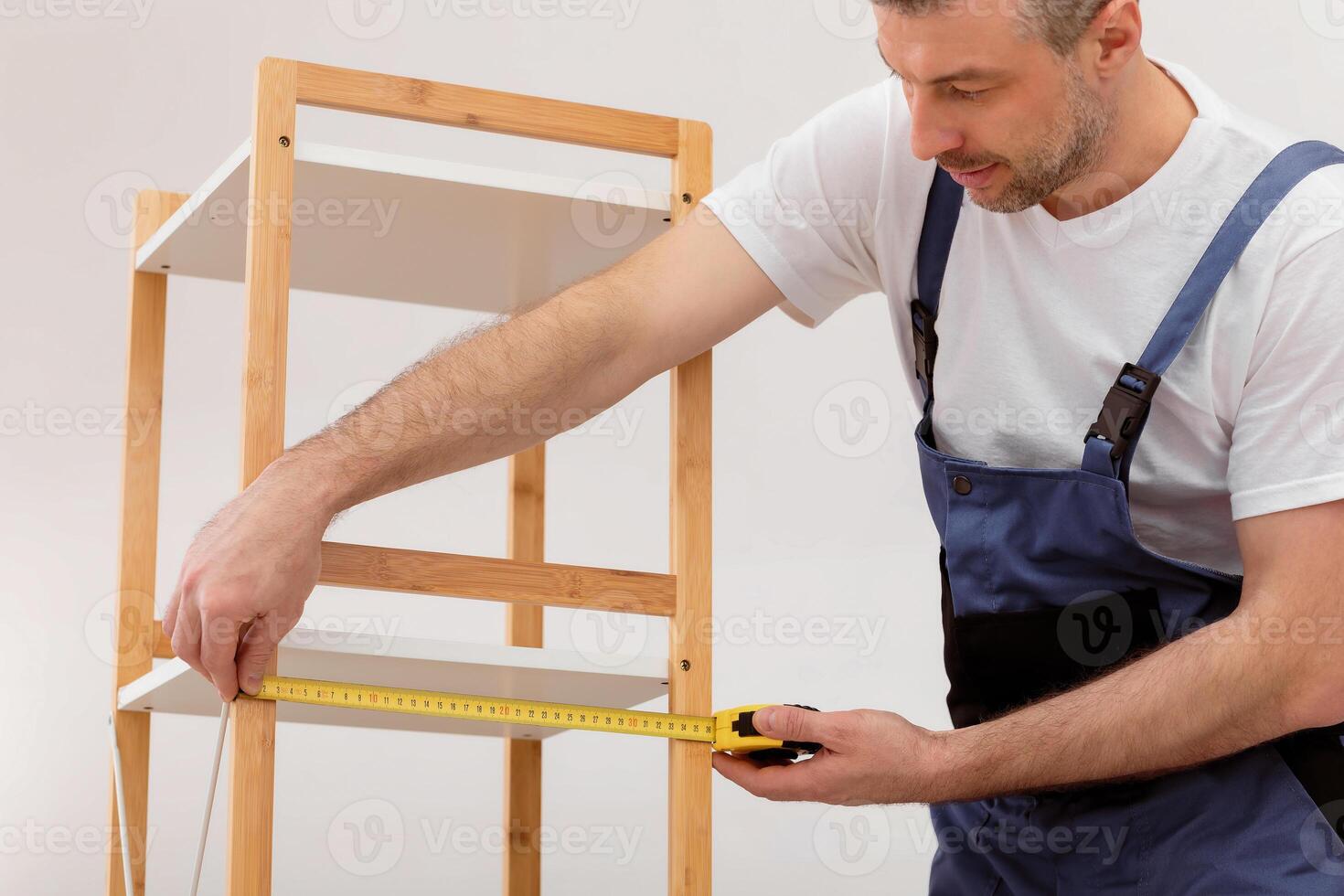 Carpentry Concept. Carpenter Making Furniture Measuring Wooden Shelf Working Indoors, Wearing Blue Uniform. Cropped photo