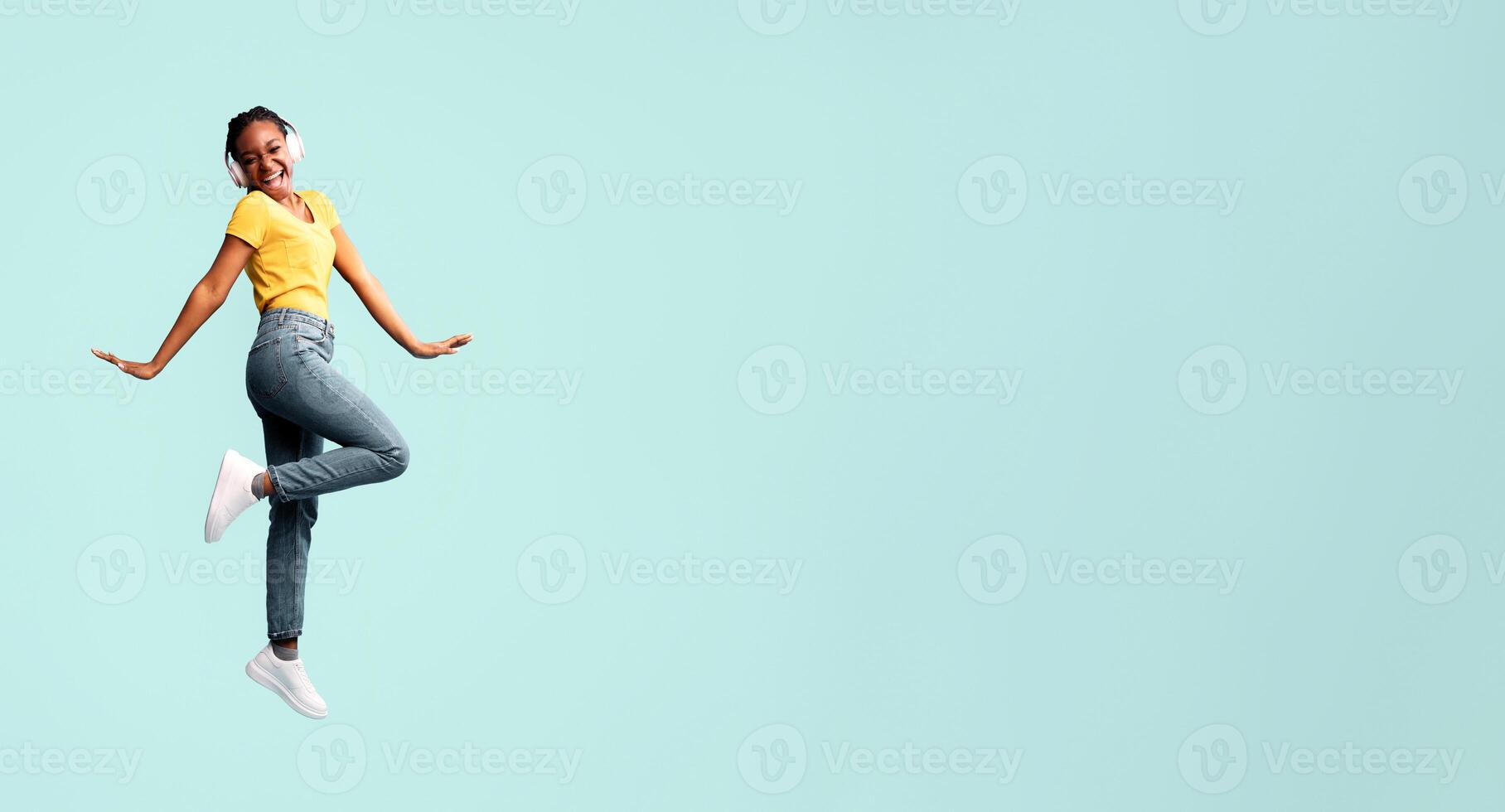 Positive African American Lady In Earphones Jumping And Singing Song Having Fun On Blue Studio Background. Empty Space For Text photo