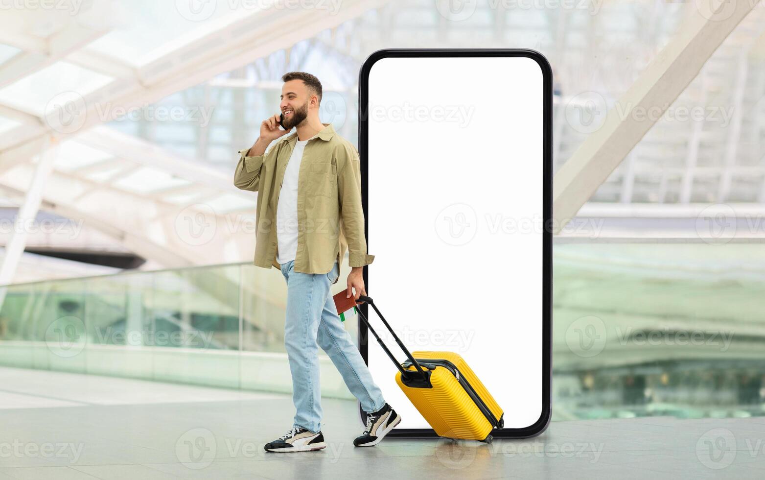 A traveler stands in a contemporary airport terminal, pulling a yellow suitcase while engaged in a phone conversation, passing by huge phone with blank screen photo