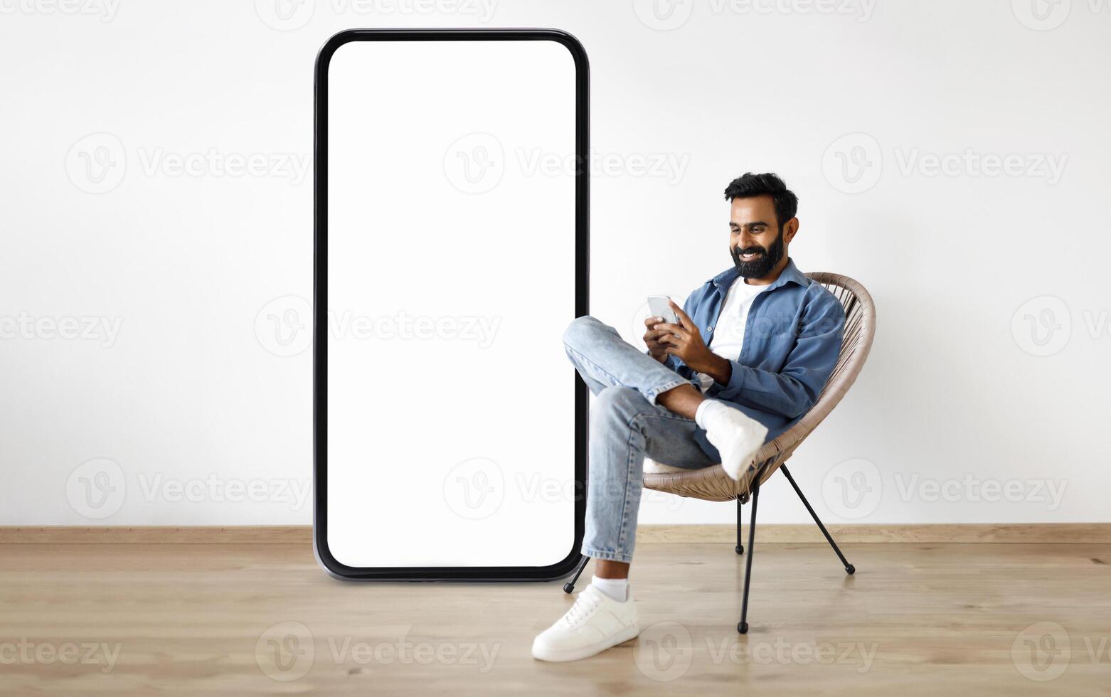 A man sits comfortably in a chair behind large smartphone with blank screen while using his smartphone, surrounded by a modern interior design that emphasizes relaxation and technology. photo