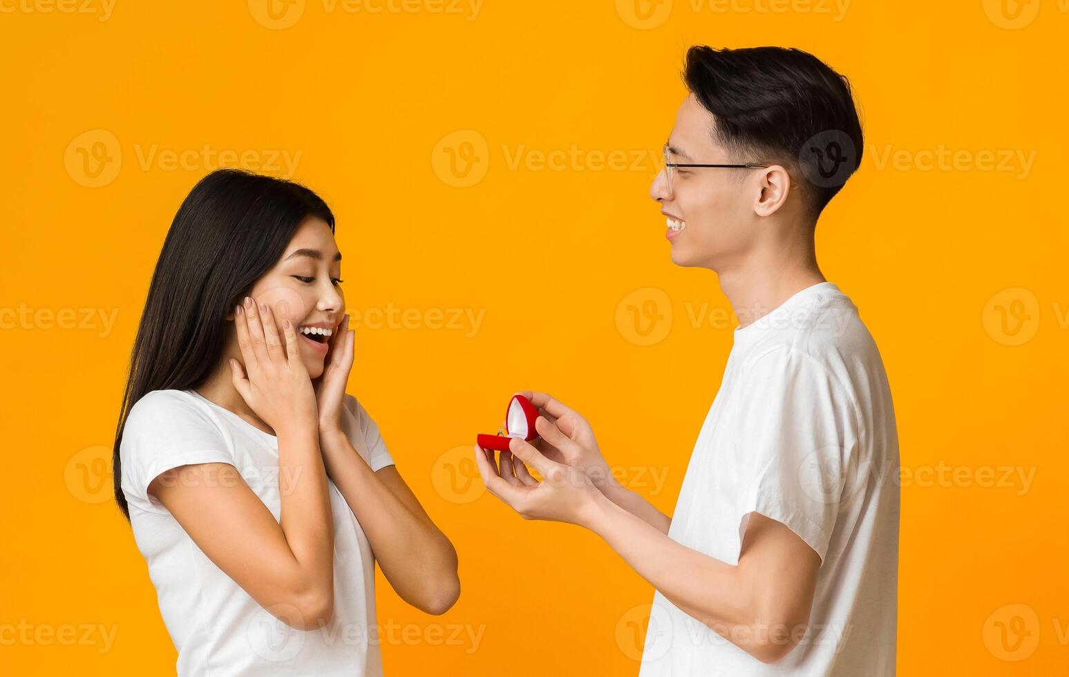 Asian young man in love making proposal to his amazed girlfriend, orange background photo