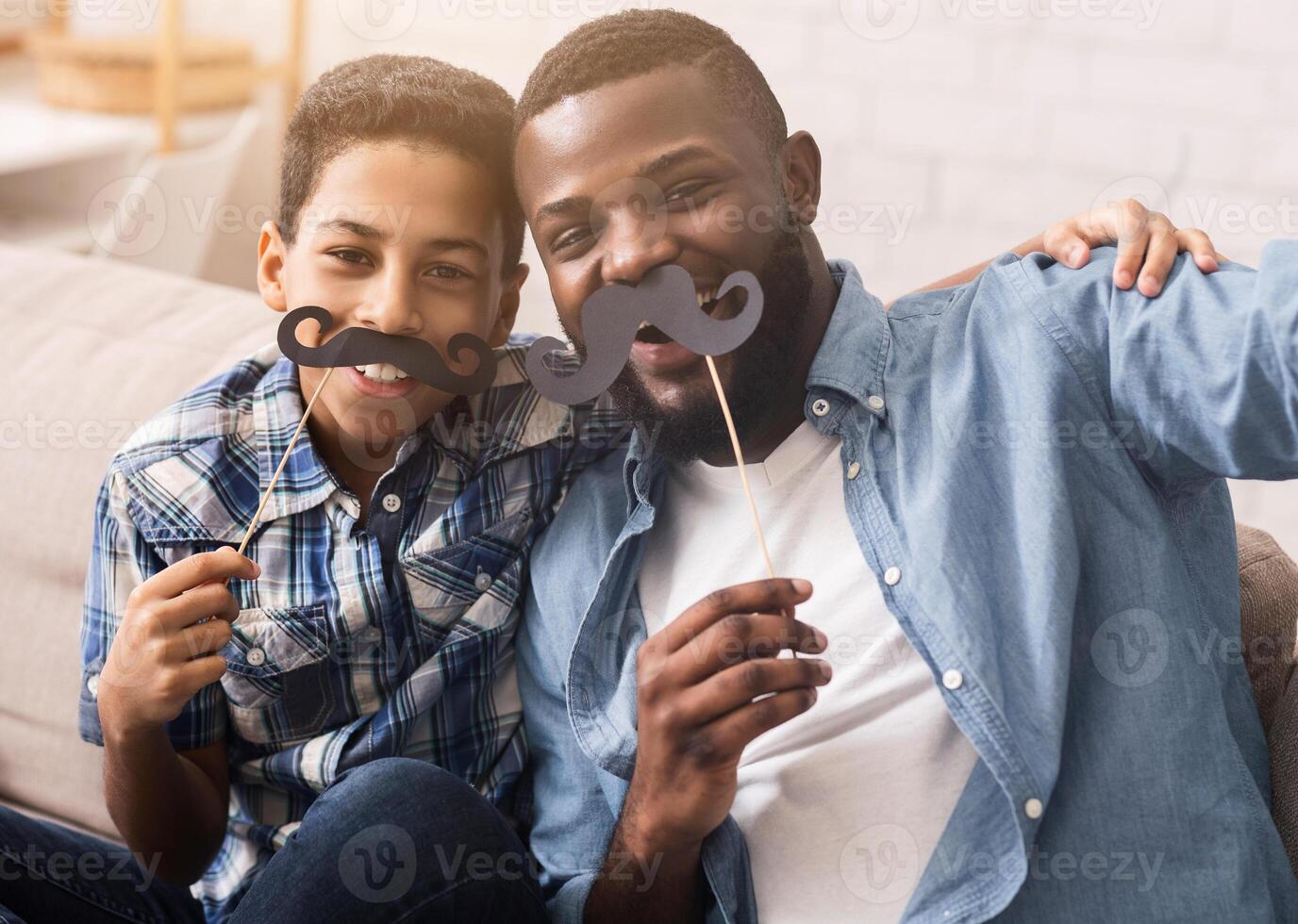 Black father and his preteen son taking selfie, holding fake moustache on sticks and smiling at camera, having fun at home photo