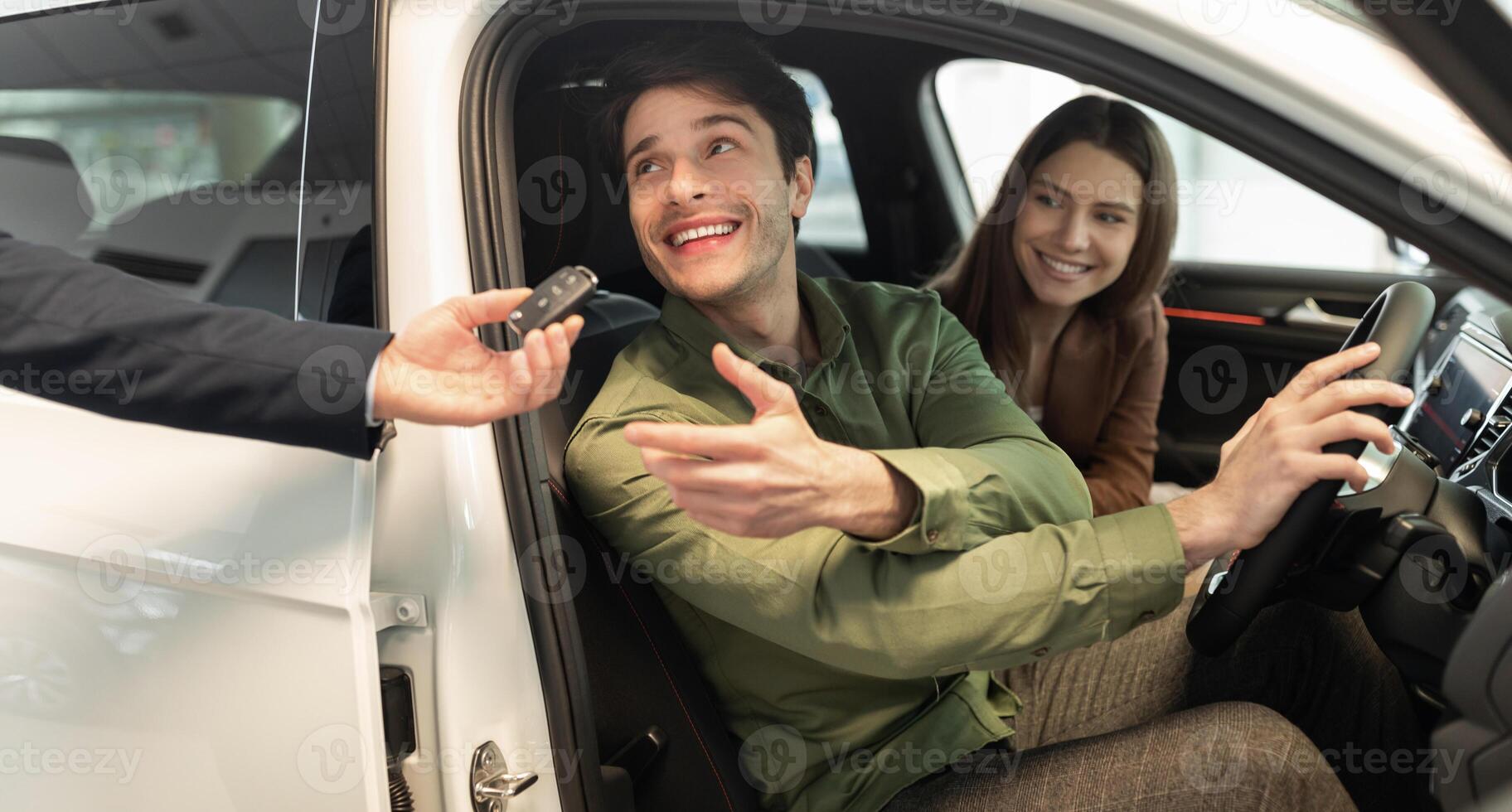 Happy millennial couple taking car key from auto salesman, sitting inside modern automobile at dealership, panorama. Cheery young family buying new vehicle at modern showroom photo