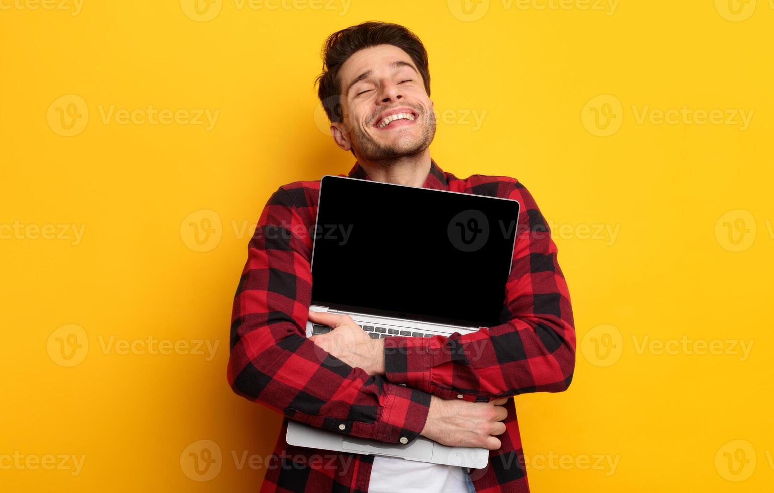Technology Lover Concept. Happy man hugging laptop with black blank empty screen, holding it tight near chest. Casual male geek with closed eyes standing isolated on yellow orange studio wall photo