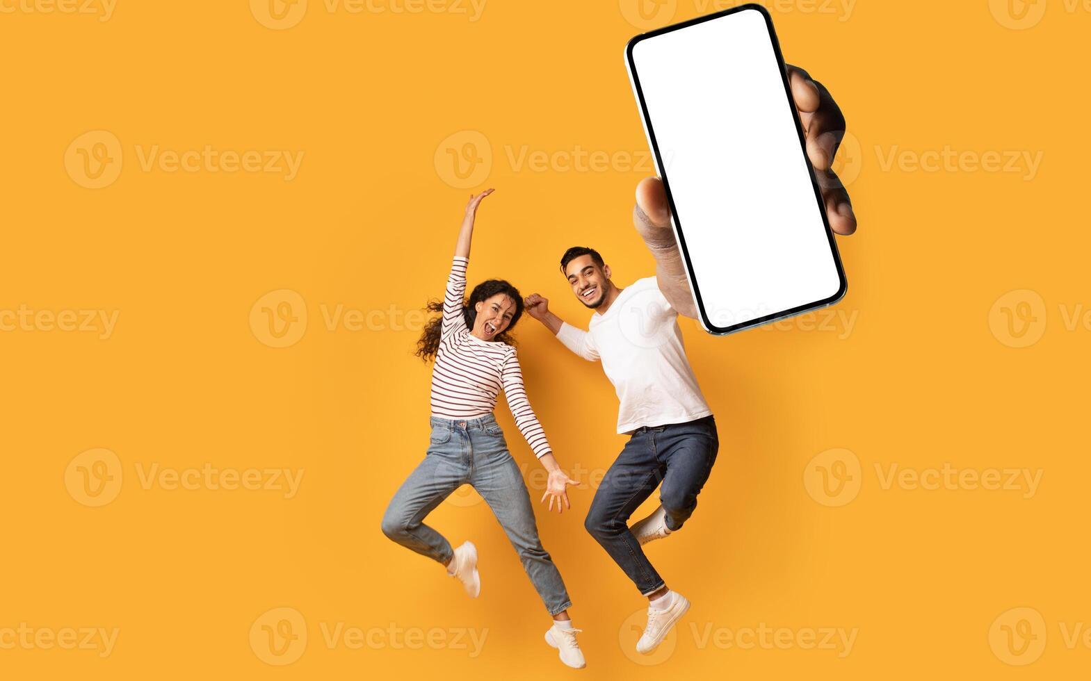 Wow, great news. Overjoyed couple showing huge smartphone with blank white screen, jumping and celebrating victory over orange studio background, mockup collage photo