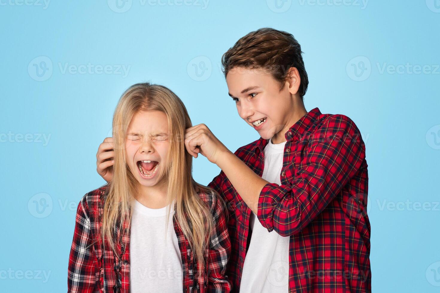 Sibling relationship concept. Elder brother teasing little sister, pulling her ears till she scream, standing together over blue studio background photo