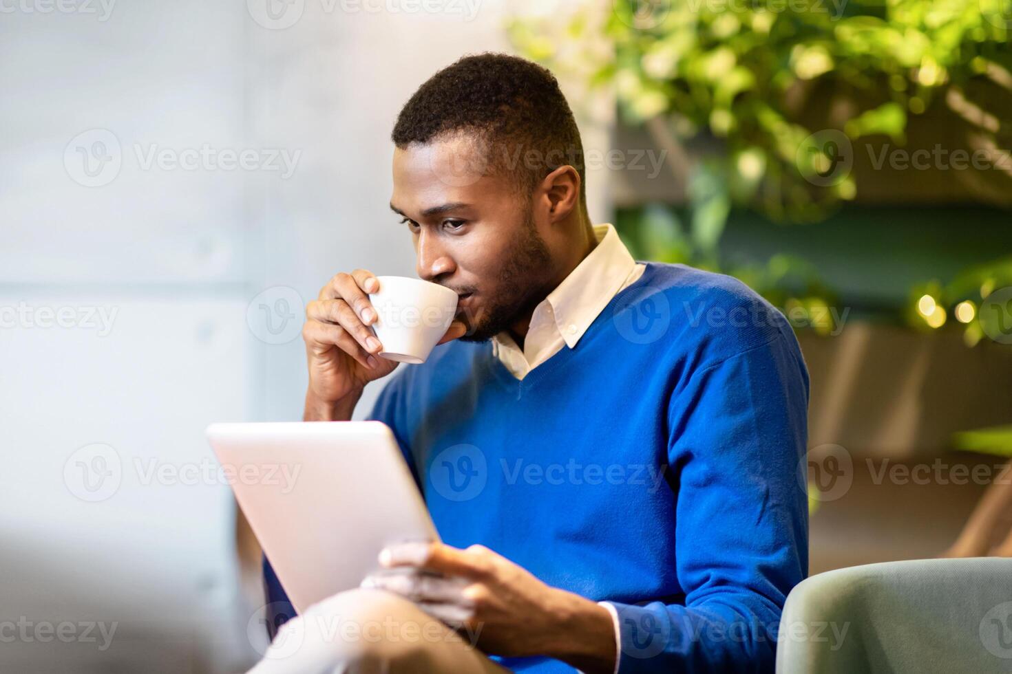 Reading News. Happy afro man using digital tablet computer and drinking coffee sitting in modern cafe, copyspace photo