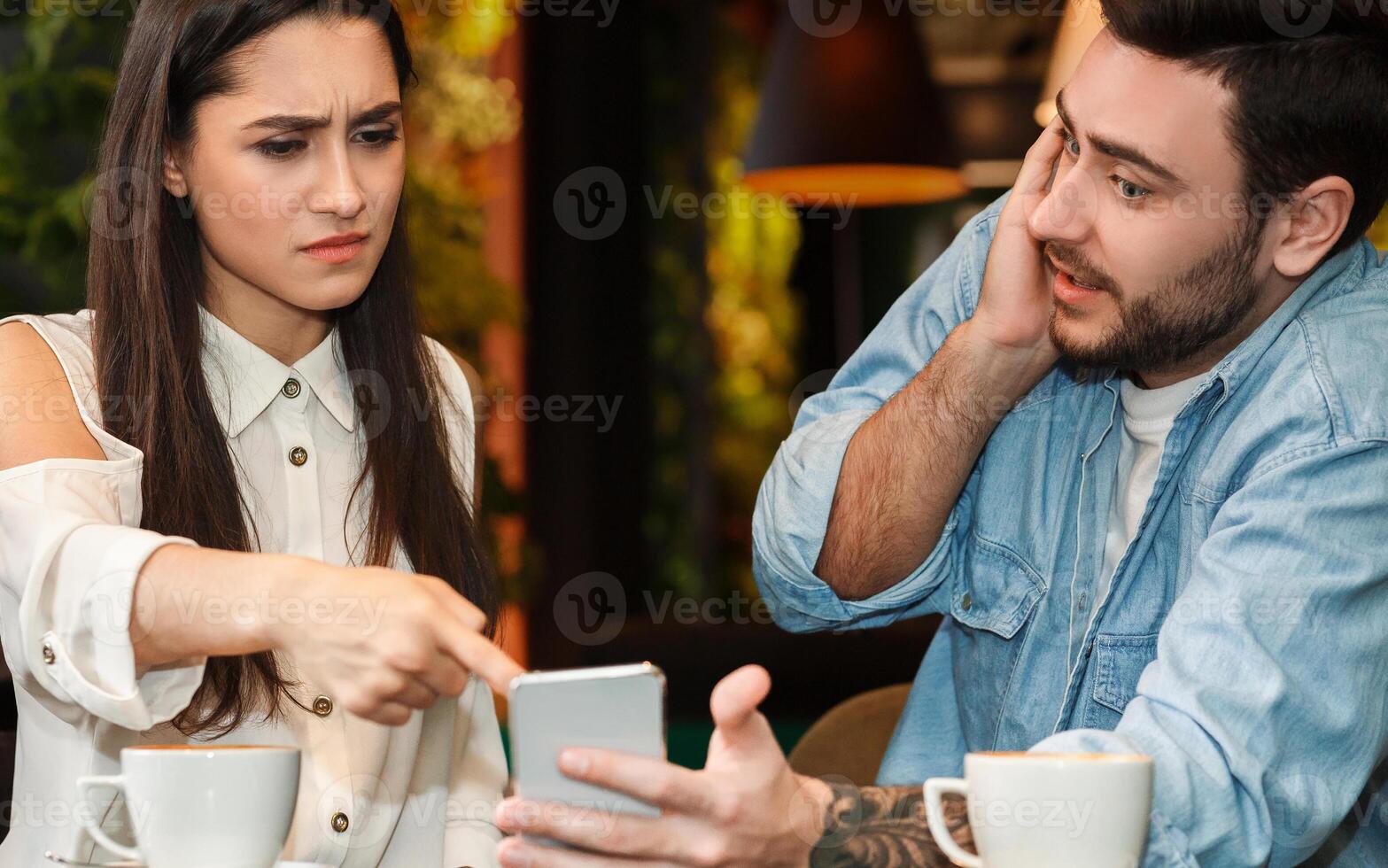 Infidelity Concept. Jealous Girlfriend Pointing Finger At Cheating Boyfriend's Phone Demanding Explanation Sitting In Cafeteria. Selective Focus photo