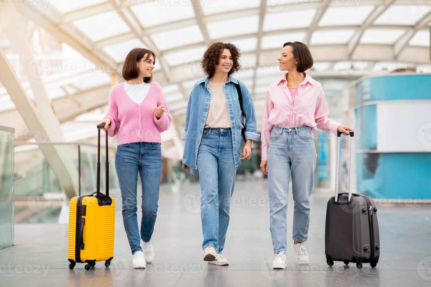 Three friends walk together through an airport terminal, each pulling a suitcase. They are engaged in conversation, smiling, and radiating excitement about their trip. photo