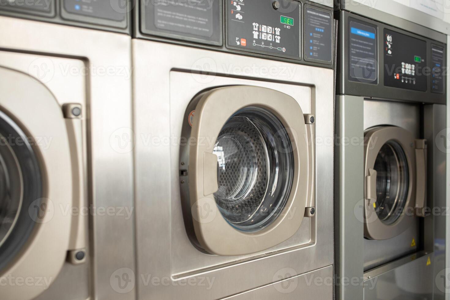 Three stainless steel washing machines are lined up in a laundromat, showcasing their circular glass doors. The bright interior highlights the machines in use during the day. photo