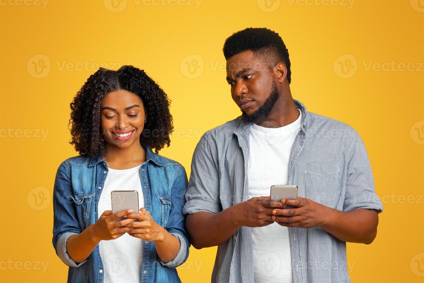 Jealous black guy spying oh his girlfriend's phone, trying to read her messages, standing together over yellow background in studio, free space photo