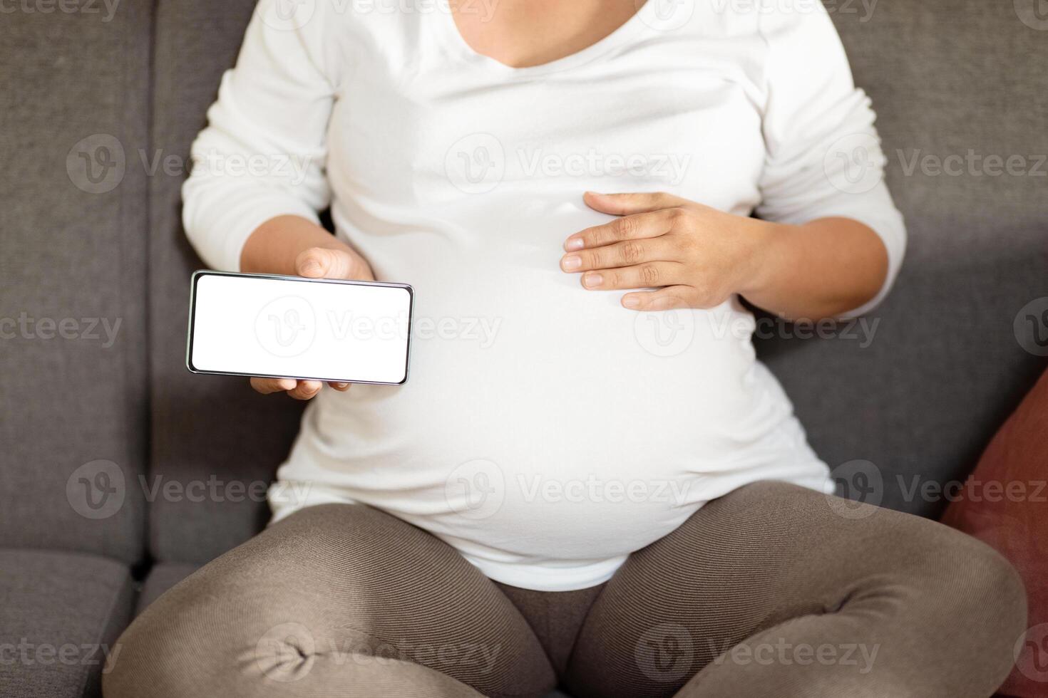 A pregnant woman relaxes on a couch, gently cradling her belly with one hand while holding her phone with the other. The atmosphere is calm, suggesting a cozy afternoon moment. photo
