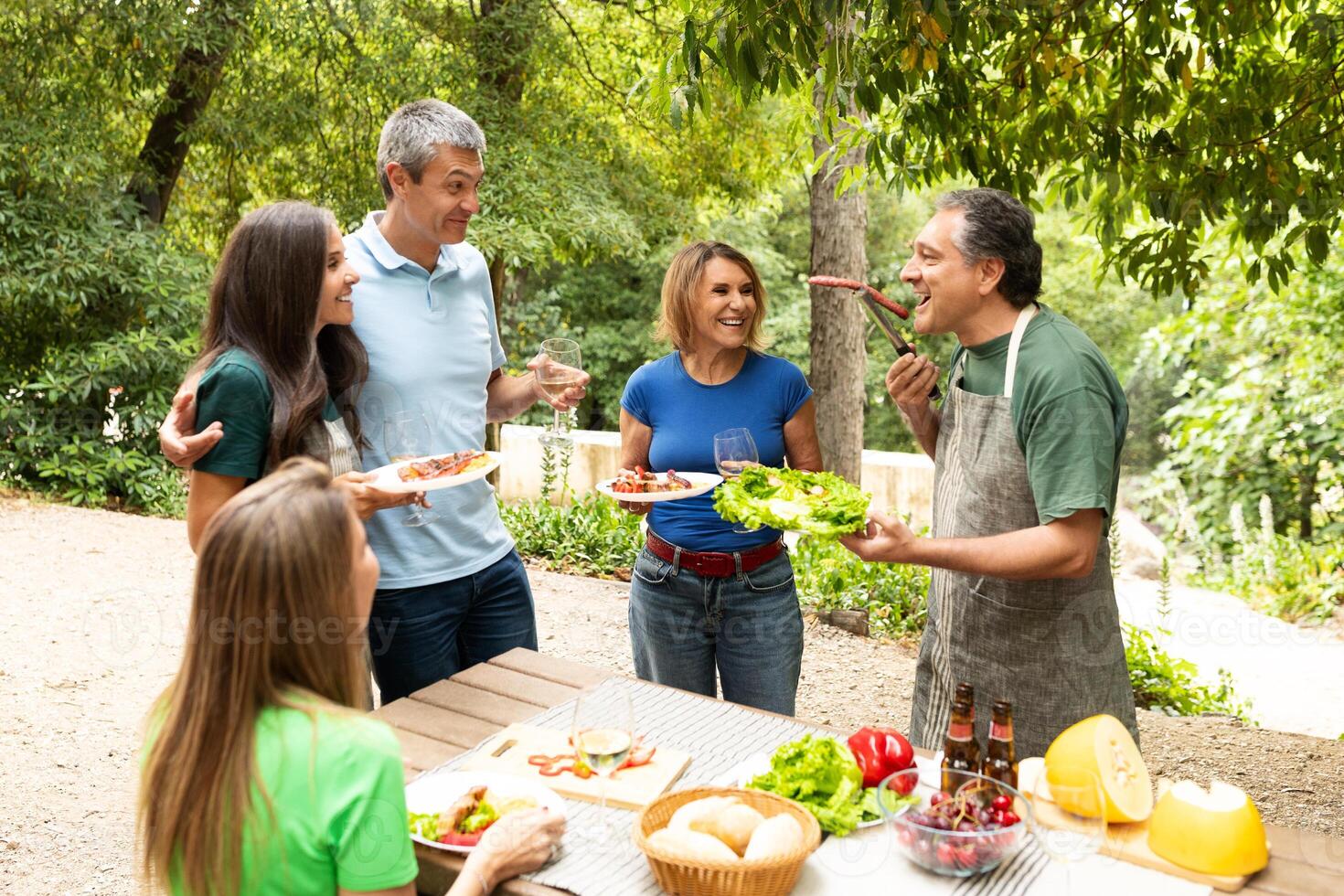 A lively group of friends shares joyful moments during a barbecue in a scenic park, indulging in delicious food and drinks while enjoying each other company in perfect weather. photo