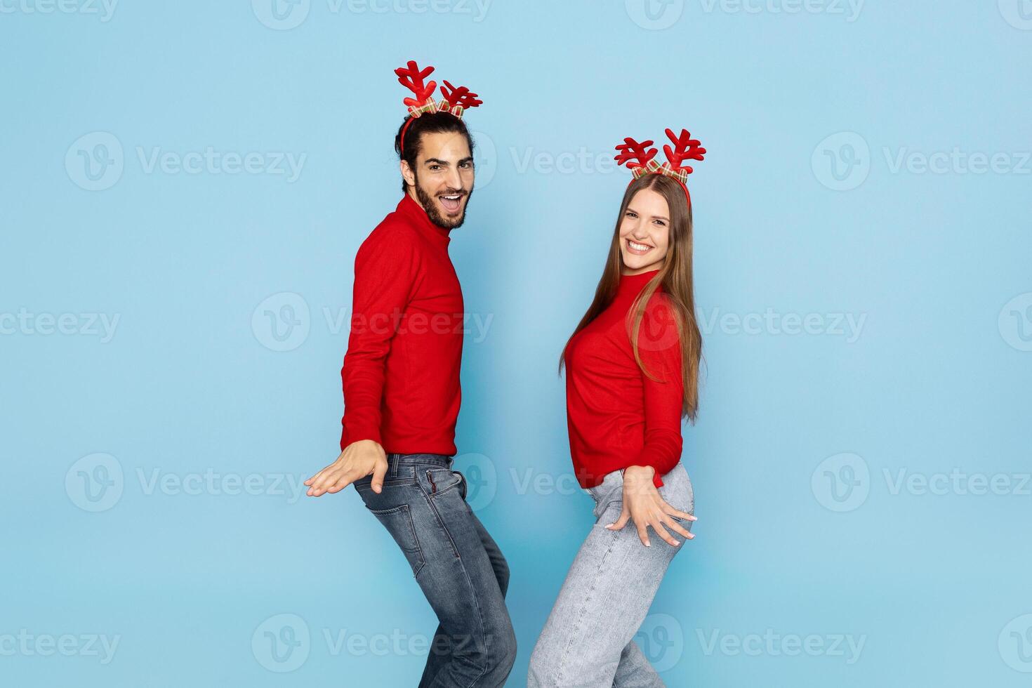 A cheerful couple in matching red sweaters and reindeer antlers poses playfully against a light blue background, exuding holiday spirit and joy during the festive season. photo