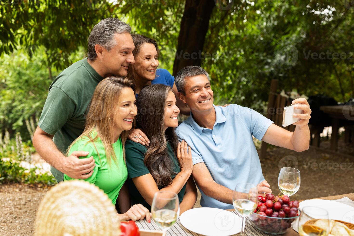 A cheerful group of five adults gather around a table outdoors, smiling and taking a selfie amidst greenery. They are enjoying a meal with grapes and drinks, creating joyful memories. photo
