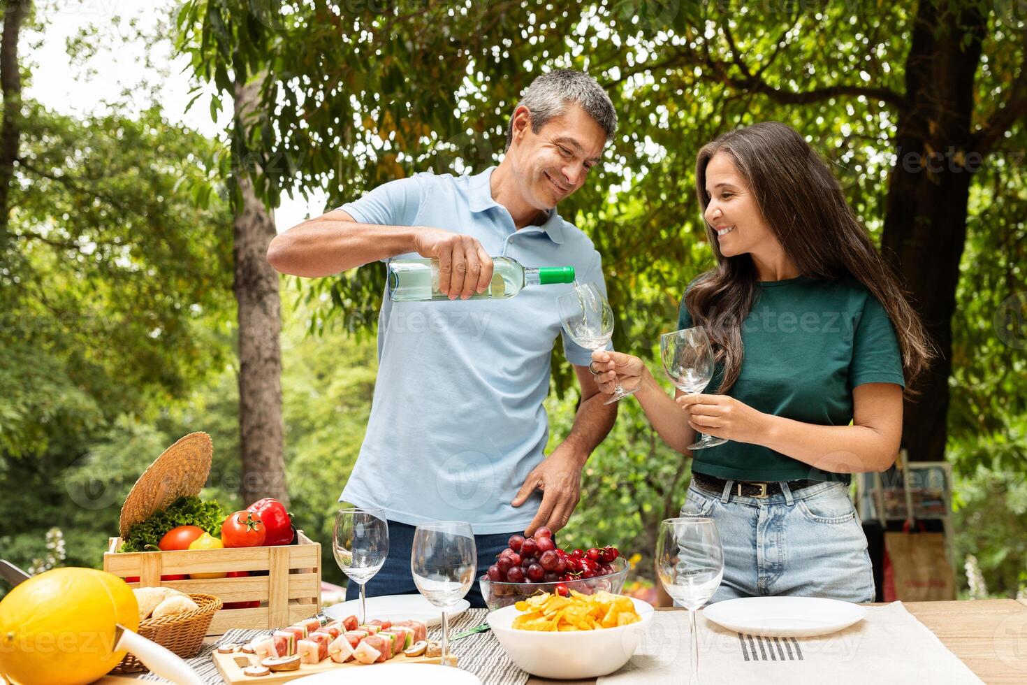 A man pours wine for a woman during a joyful summer picnic in a lush green park, surrounded by fresh fruits and a variety of snacks, creating a festive and relaxed atmosphere. photo