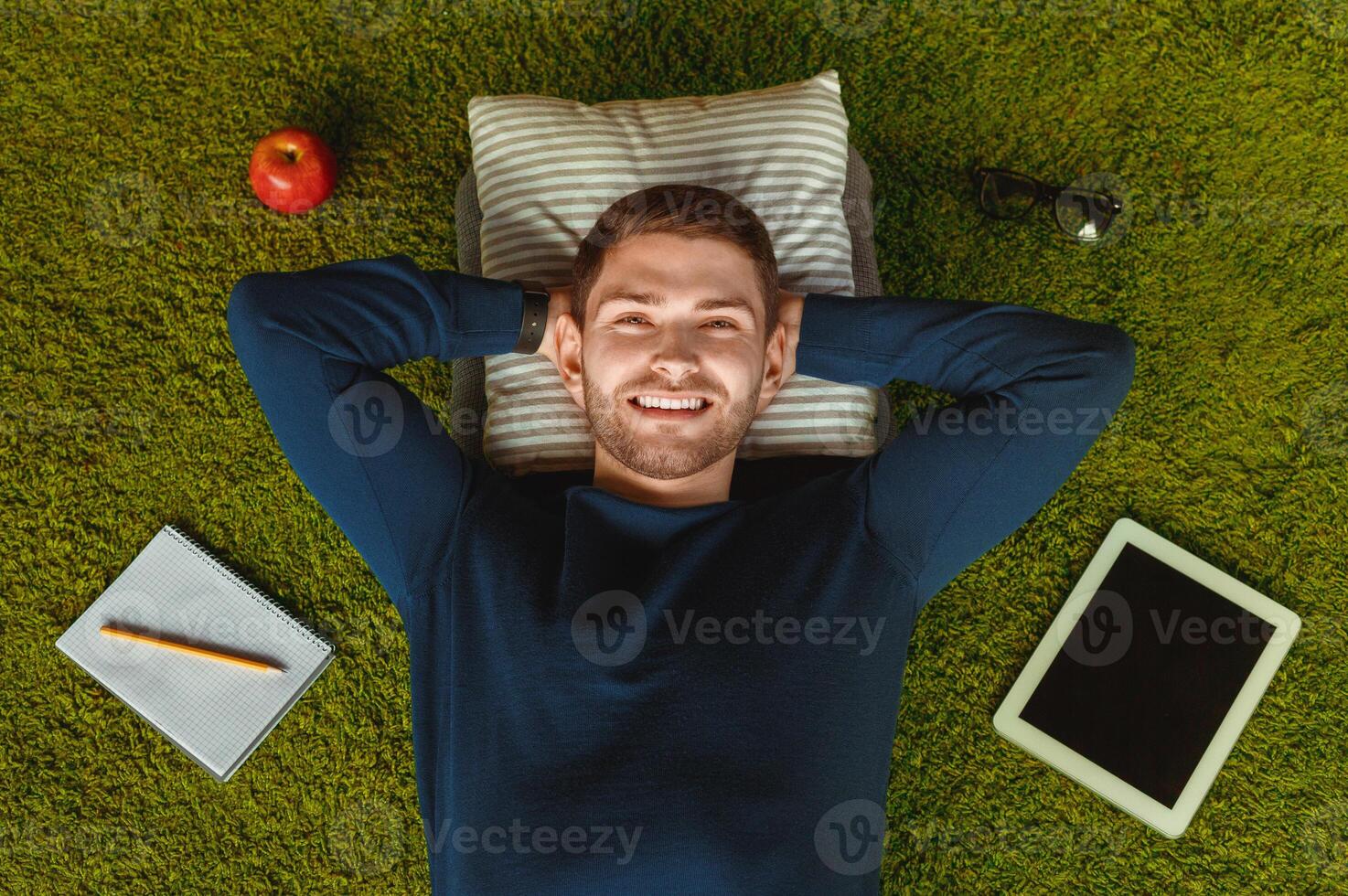 Student Lifestyle. Relaxed Guy Lying On Green Grass Carpet Among Digital Tablet, Notebook And Eyeglasses. Top View photo