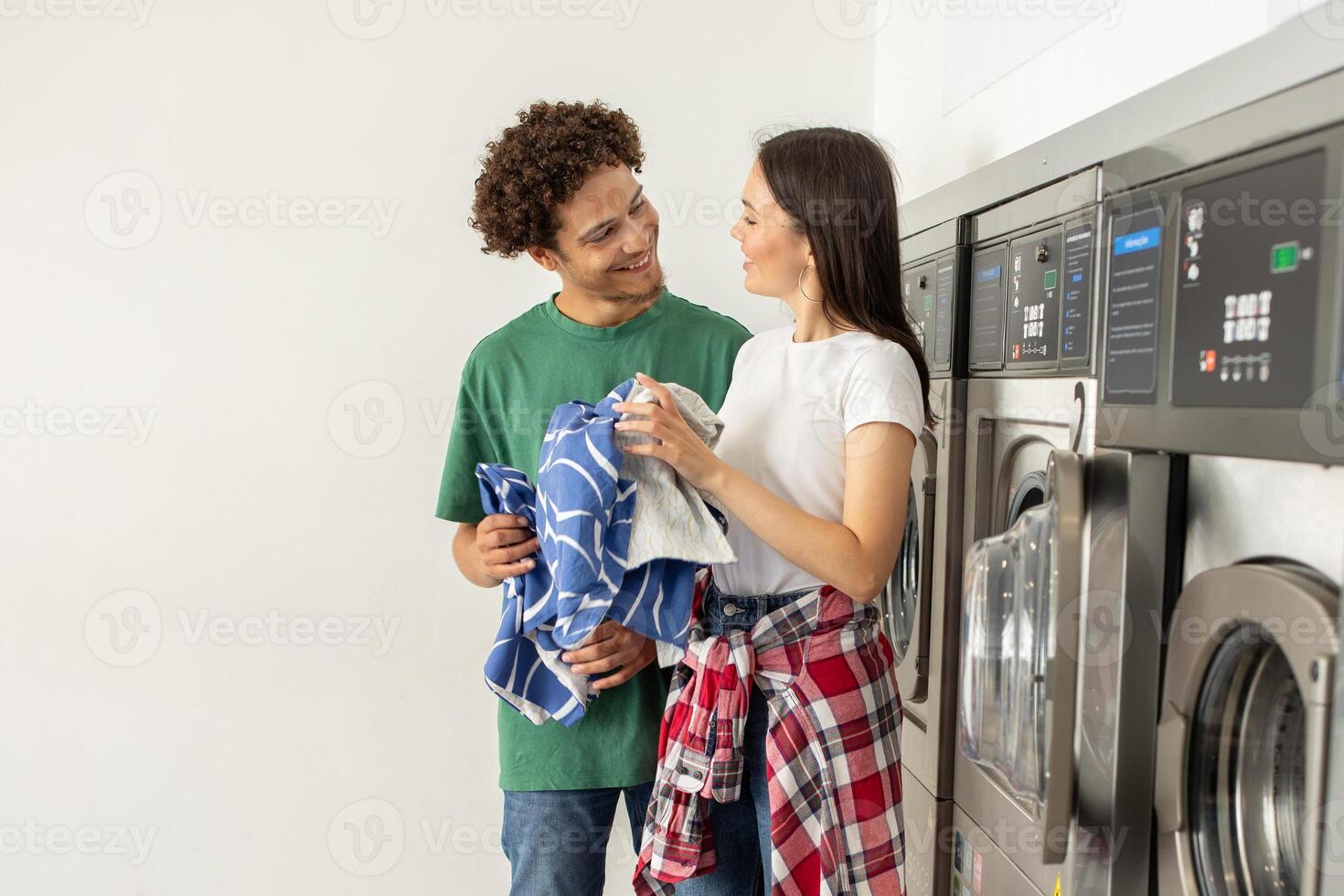 A young couple smiles at each other while holding laundry baskets in a clean and bright laundromat. They appear to enjoy their time together as they wait for their clothes to wash. photo