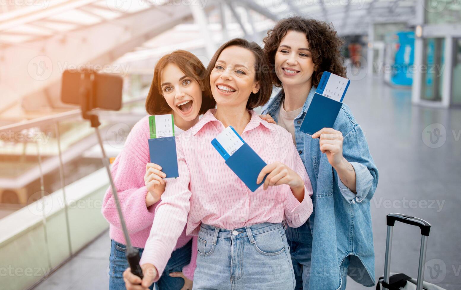 Three enthusiastic friends stand together at the airport, holding their tickets, smiling, and capturing the moment with a selfie stick. Their excitement for the upcoming journey is evident. photo