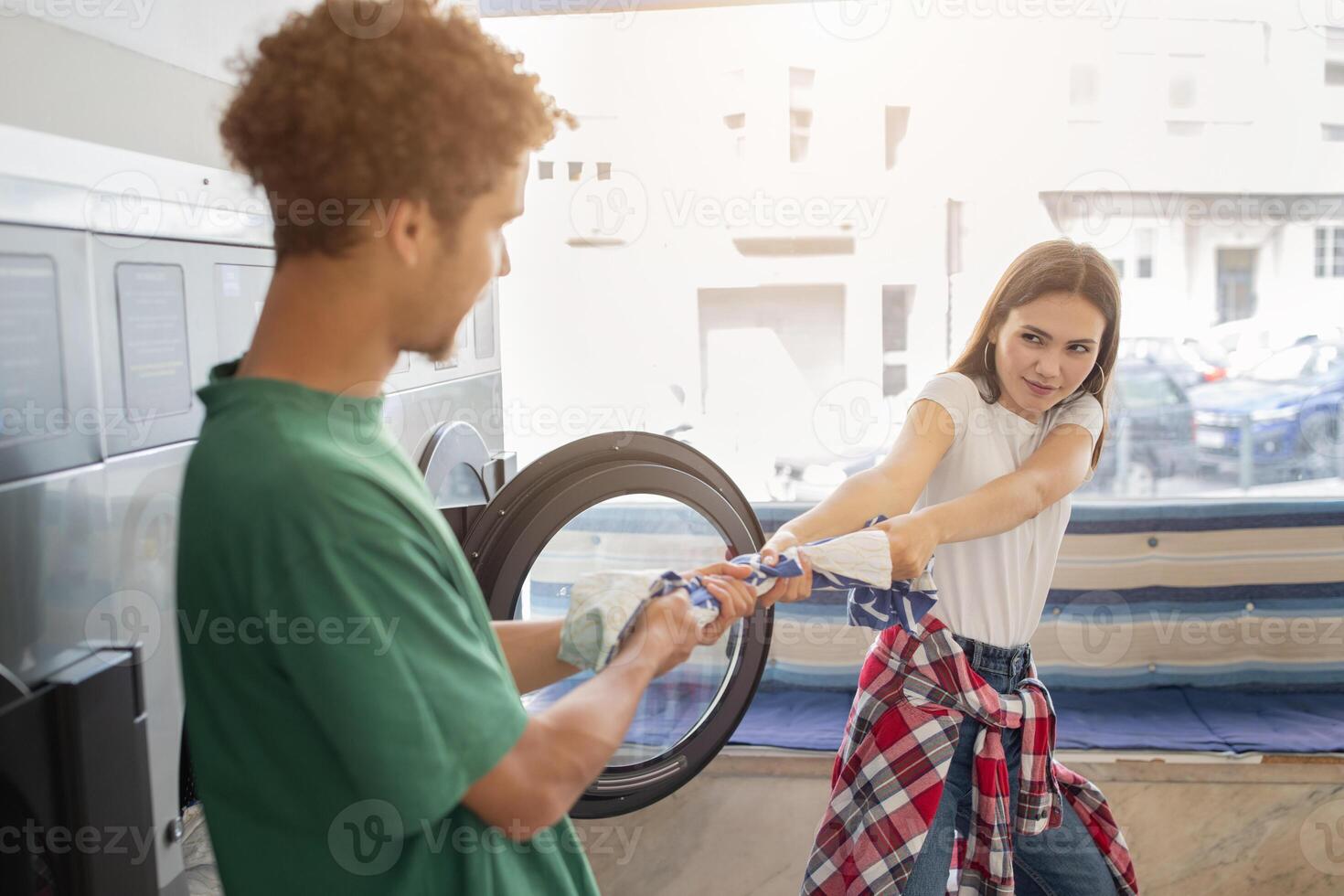 In a sunny laundromat, a man and woman engage in a lighthearted struggle over a laundry basket, showcasing playful energy amid washing machines and bright surroundings. photo