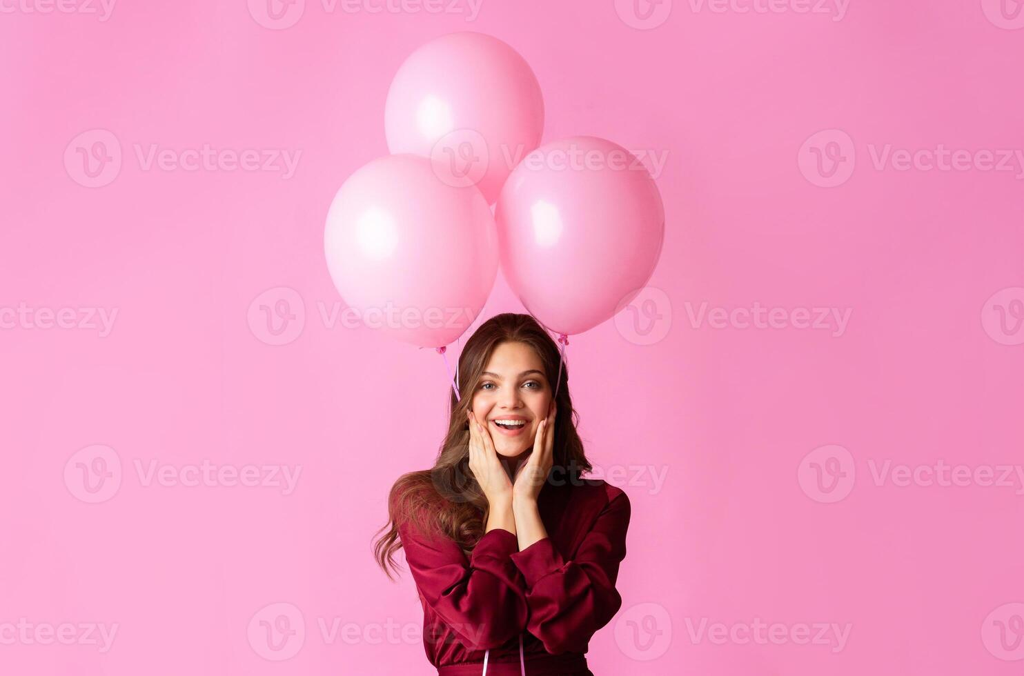 Playful Mood. Cheerful Young Lady Holding Three Rosy Balloons And Touching Cheeks In Excitement, Posing Over Colorful Background photo