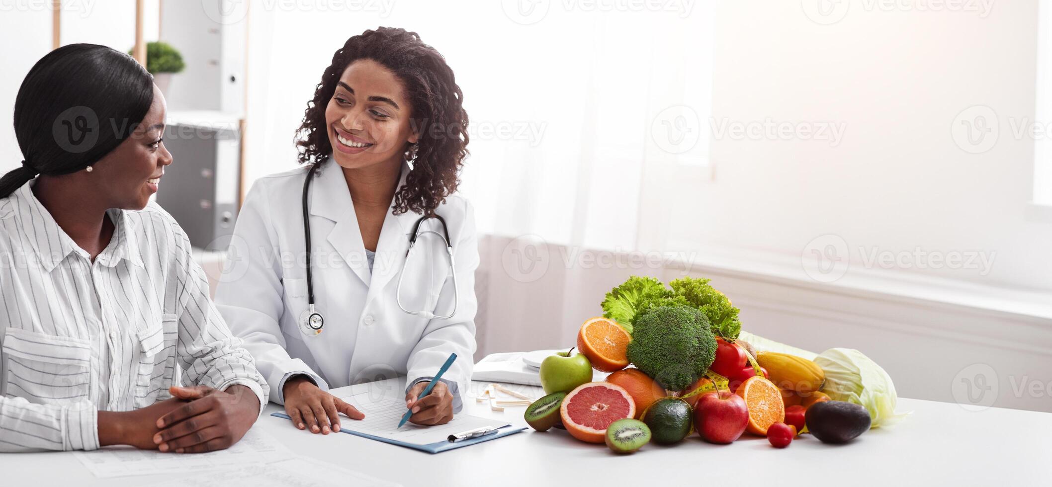 Friendly afro lady nutritionist making female patient anamnesis at clinic, discussing treatment, copy space photo