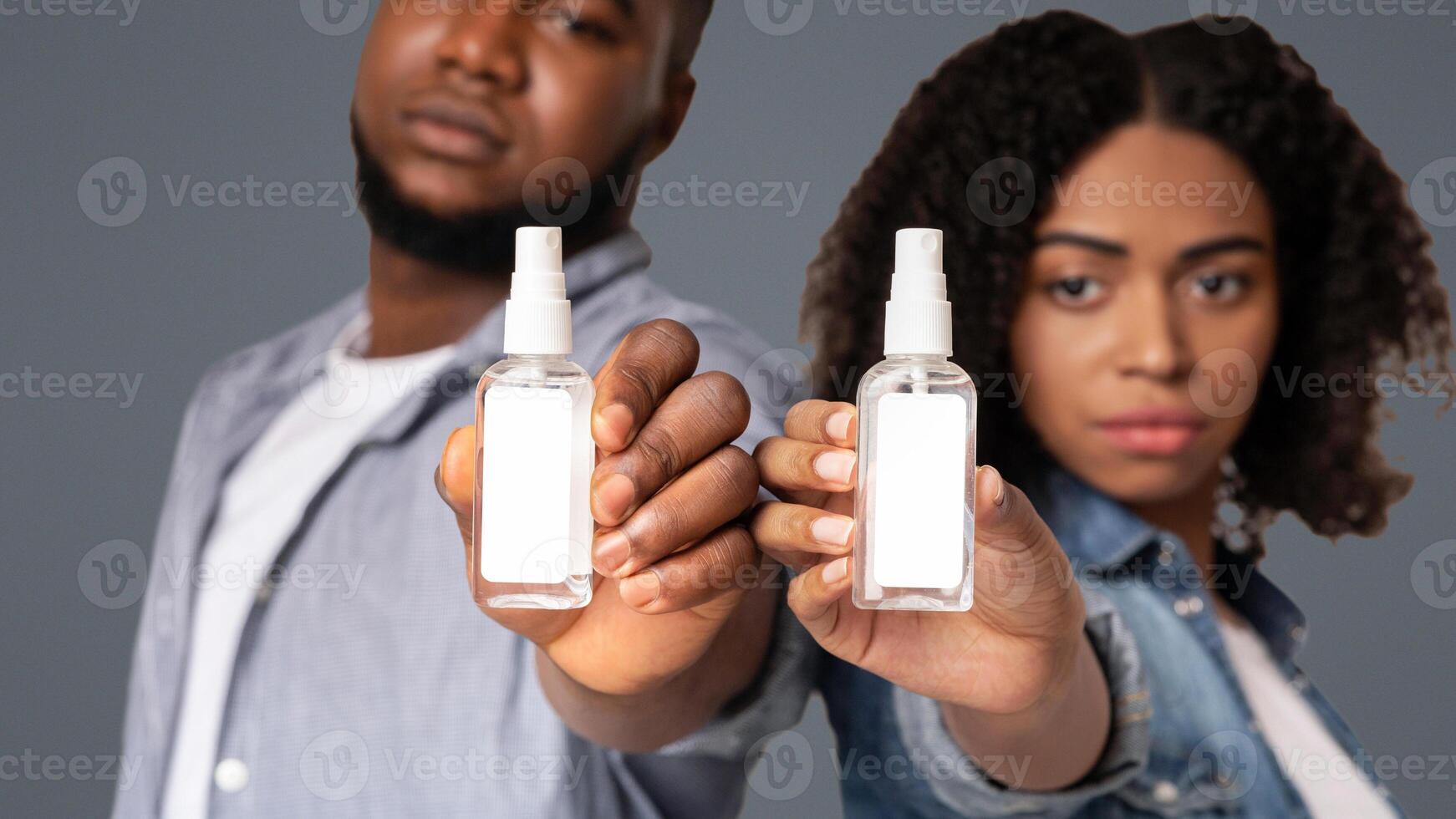 Bottles Of Disinfectant Sprays With White Sticker Label In Hands Of Young African American Couple Standing On Light Background, Selective Focus photo