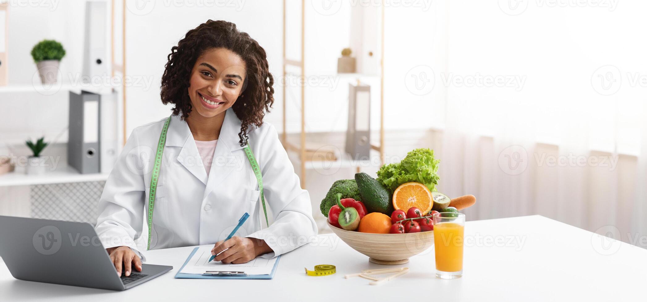 Cheerful afro woman dietologist working with laptop in modern office, having fresh juice, fruits and vegetables on table photo