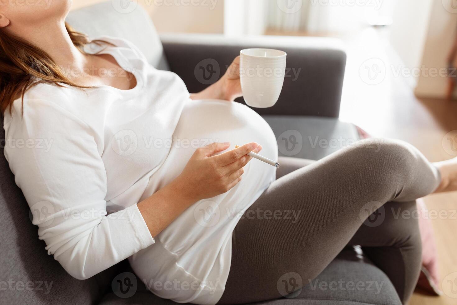 A pregnant woman wearing a white shirt and grey pants sits on a grey couch. She holds a white mug in her right hand and a cigarette in her left. photo