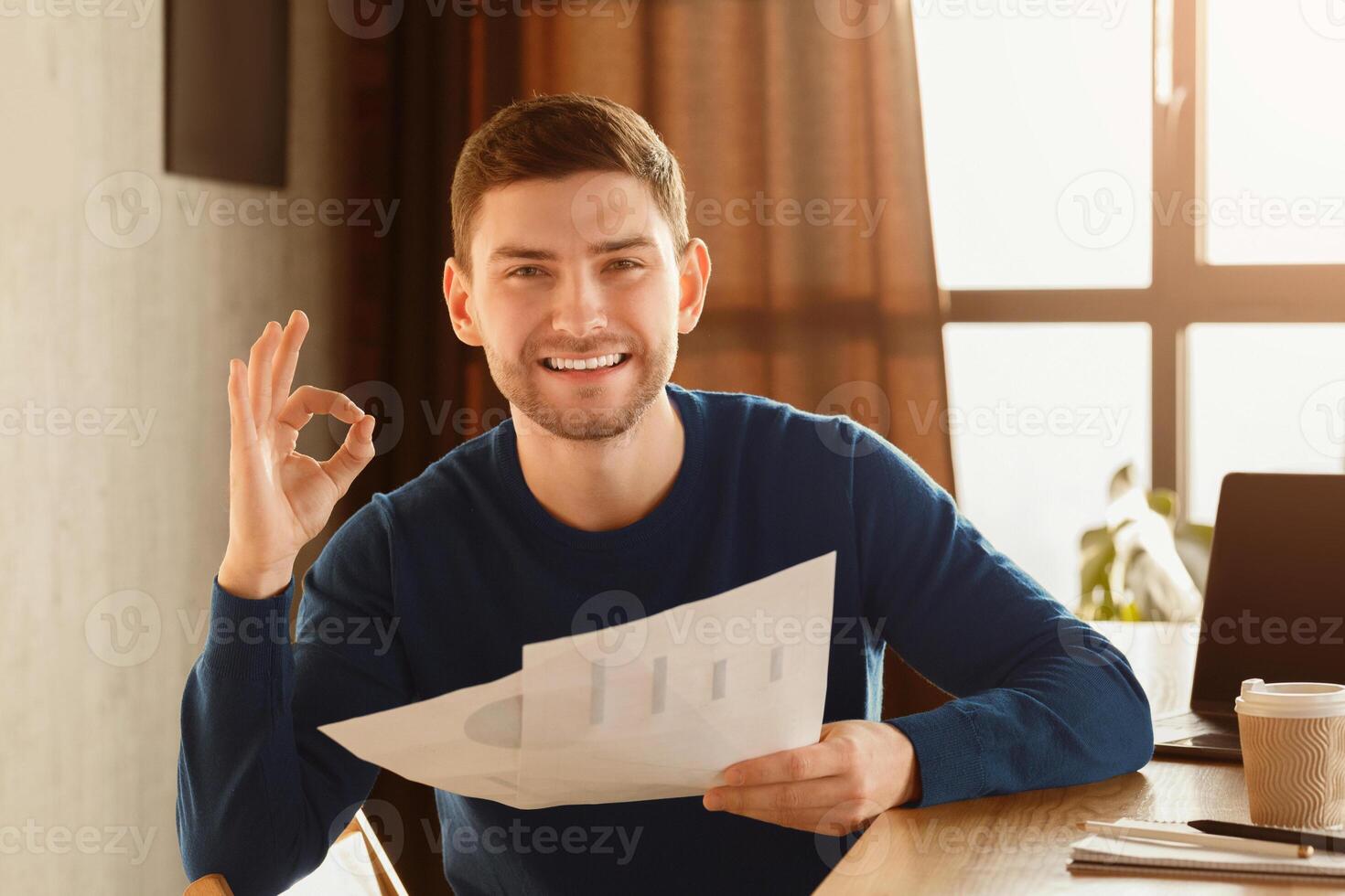 Business Startup Idea. Happy Guy Gesturing Okay Holding Papers With Graphs And Charts Sitting At Workplace Indoor. Selective Focus photo