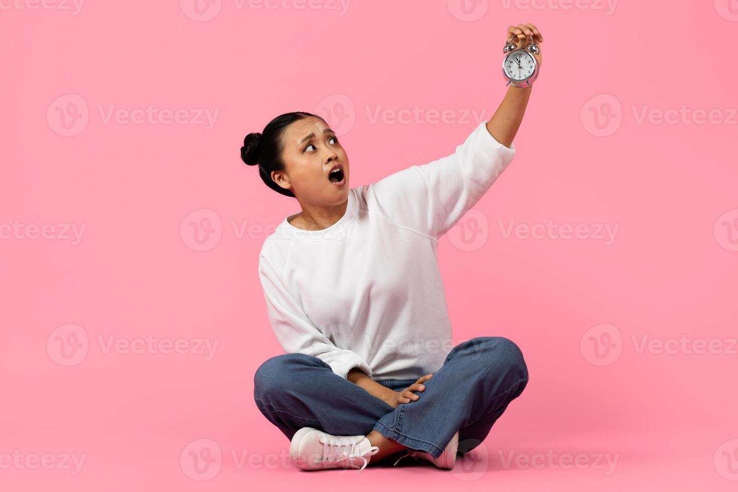 Asian woman sits on the floor with a shocked expression, raising a stopwatch in one hand against a vibrant pink backdrop. She appears startled as she looks at the time. photo