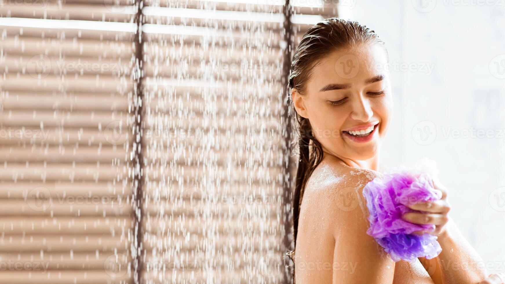 Woman washing with shower gel, standing under water drops and rubbing body with sponge, panorama with copy space photo