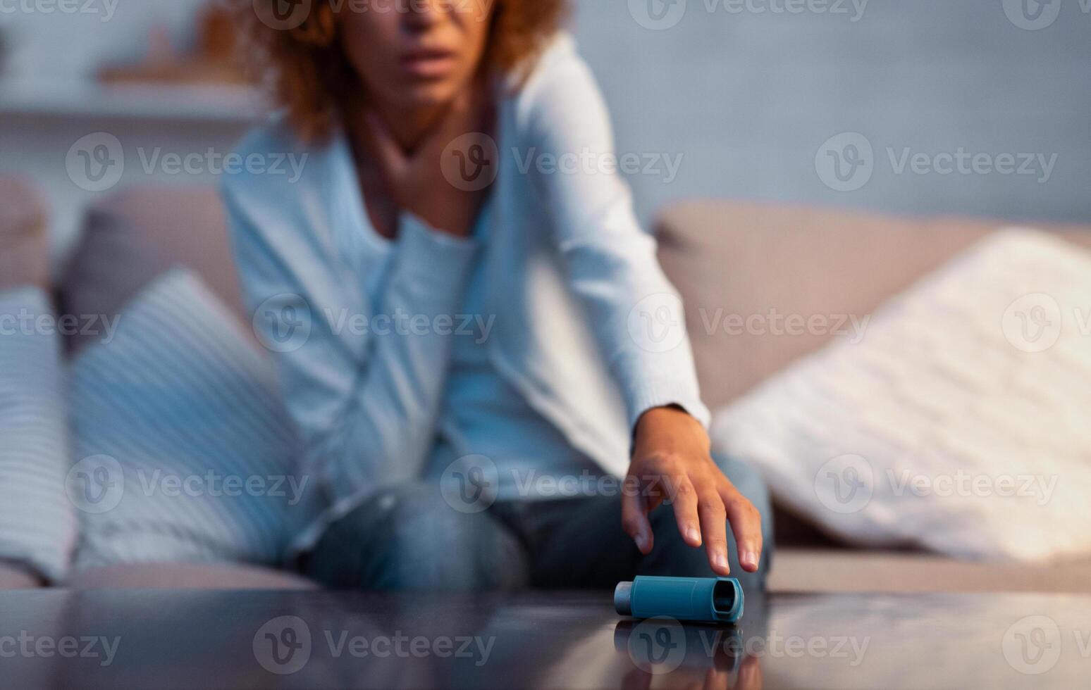Afro girl suffering from asthma and taking inhaler from table, sitting on couch in living room photo