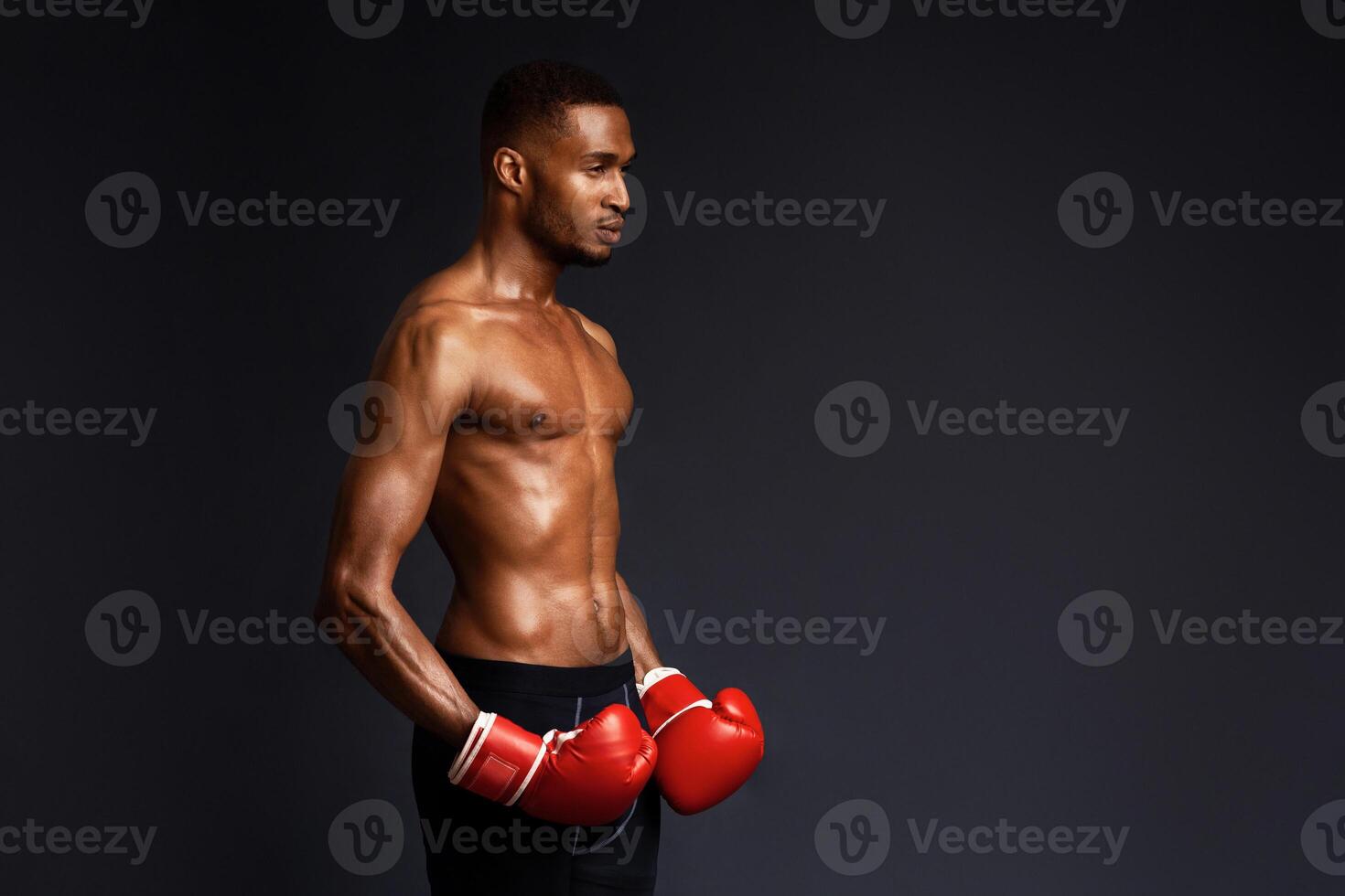 Fighter. Half-turned athletic black man in boxing gloves looking aside at copyspace isolated over black studio wall photo